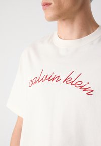 White cotton t-shirt featuring red embroidered "calvin klein" logo. Crew neckline and short sleeves. Smooth texture with a relaxed fit.