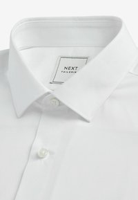 Camisa blanca de botones con cuello clásico, hecha de tela suave. Presenta un cierre de un solo botón y una etiqueta con 'NEXT TAILORING.'