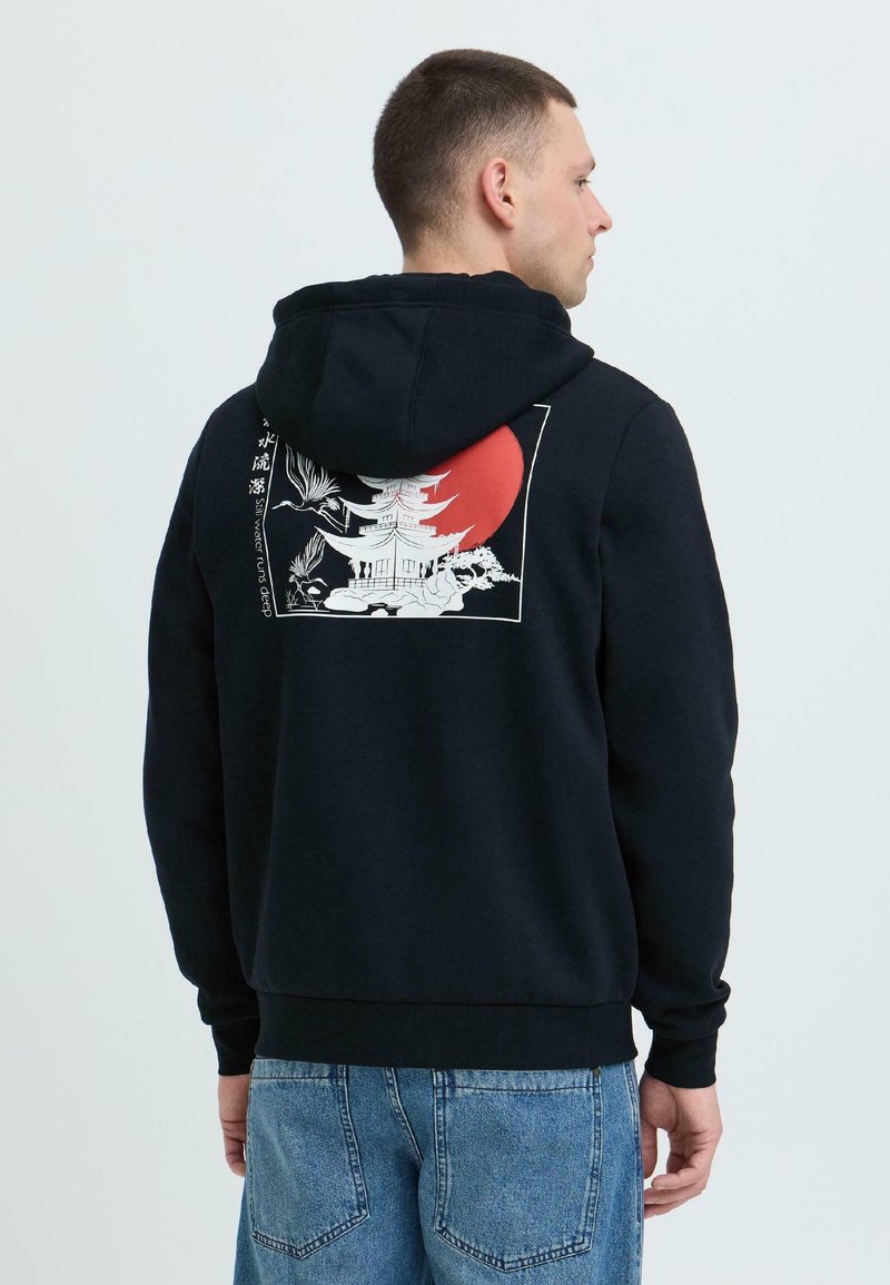 Homme portant un sweat à capuche noir avec la capuche relevée et un motif au dos représentant une pagode blanche, des grues en vol et un grand soleil rouge.