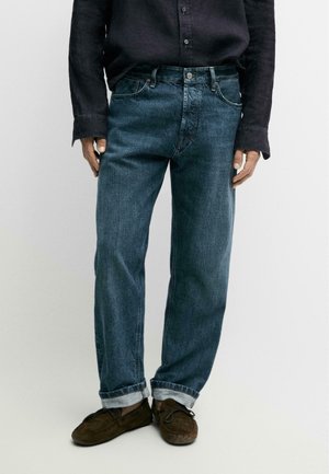 Man draagt donkerblauwe spijkerbroek met opgerolde pijpen, donker shirt met lange mouwen en bruine suède mocassinachtige schoenen, staand tegen een effen achtergrond.