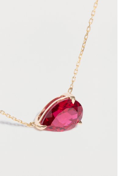 Collar de cadena de oro con un colgante grande en forma de lágrima con una piedra preciosa roja, montado en un broche de oro sobre un fondo blanco.