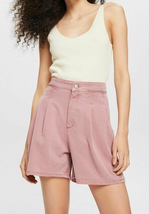 Femme portant un haut crème sans manches côtelé et un short ample rose taille haute, debout devant un arrière-plan uni.