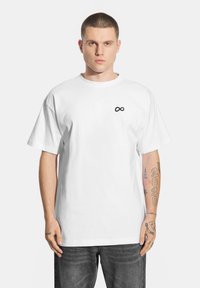 Biała bawełniana koszulka, luźny krój, z małym czarnym logo symbolu nieskończoności na lewej piersi. Noszona z ciemnymi dżinsami.