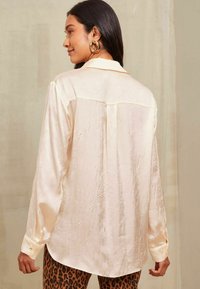 Cămașă din satin crem, cu mâneci lungi și o textură netedă, un model subtil de cute și cu guler. Prezintă manșete cu nasturi și detalii la spate.