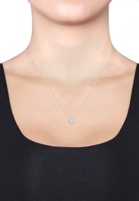 Zilveren halsketting met een fijne ketting, voorzien van een ronde hanger versierd met kleine, glinsterende stenen, rustend op een bloot decolleté.