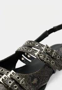 Μαύρα και γκρι σανδάλια με μοτίβο ερπετού, με πολλαπλούς ιμάντες που διαθέτουν ασημένια εξαρτήματα και διακοσμητικά μεταλλικά κουμπιά σε λεπτή σόλα.