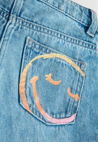 Blå denimshorts med en baglomme med et smilende ansigtsdesign i pastelfarver, der fremhæver tekstureret stof og syede detaljer.
