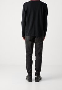 Schwarzes Langarmshirt mit rotem Kragen, kombiniert mit dunkelgrauen Jeans und schwarzen Schuhen, von hinten auf einem weißen Hintergrund gezeigt.