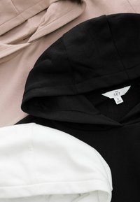 Beige Jacke, schwarzer Hoodie mit Aufschrift "THE SET S" und weißer Hoodie übereinander getragen, die Kapuzen und Stoffstrukturen hervorheben.
