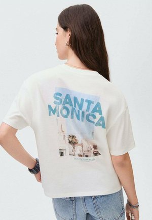 T-shirt en coton blanc avec un graphique bleu "SANTA MONICA" et une scène de plage au dos. Coupe oversize, manches courtes et décolleté décontracté.