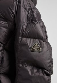 Manica di giacca trapuntata nera con patch triangolare nero e argento "JACKIT" sulla parte superiore del braccio contro uno sfondo grigio chiaro.