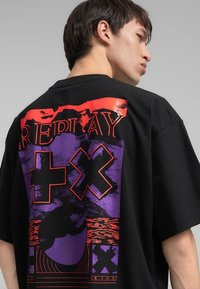 T-shirt nera oversize con un design grafico che presenta accenti rossi e viola, testo in grassetto e motivi astratti sul retro.