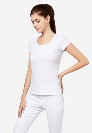 Frau mit langen braunen Haaren im Pferdeschwanz trägt ein schlichtes weißes Kurzarm-T-Shirt und weiße Hosen, steht mit einer Hand in der Hüfte.