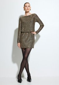 Robe en sequins dorés métalliques avec des manches longues, un décolleté bateau et une taille froncée. Associée à des bas noirs et des escarpins noirs à bout pointu.