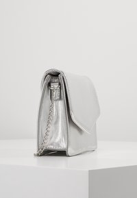 Srebrna torebka o metalicznym wykończeniu, z wyraźnie zarysowanym kształtem, z klapą, łańcuszkowym paskiem i gładką fakturą. Posiada geometryczny kształt i ostre krawędzie.