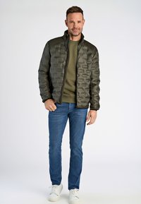 Olivgrön vadderad jacka med strukturerad yta, hög krage och dragkedja, matchad med en mörkgrön tröja och blå jeans. Vita sneakers.