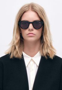 Mustat pyöreät aurinkolasit, joissa on tummat linssit, paksu kehys ja sileä pinta. Mallilla on päällä musta takki valkoisen kauluspuseron päällä.