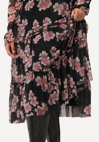 Vestido negro con tela translúcida que presenta un patrón floral en rosa y blanco. Las capas con volantes y las mangas largas añaden textura y detalle.