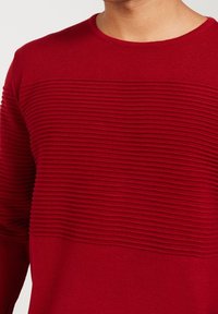 Maglione rosso con una texture a costine, caratterizzato da strisce orizzontali sul petto. Collo rotondo e maniche lunghe. Tessuto a maglia morbido.