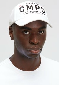 White baseball cap with black "CMPD" text and logo, featuring a curved brim and adjustable strap. Worn with a white crewneck shirt.