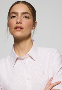 Camicia button-up a righe rosa chiaro con colletto e piccolo logo ricamato sul petto. Il tessuto appare liscio e fresco.