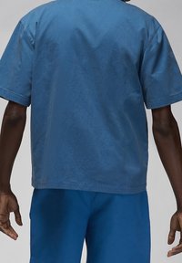 Persona che indossa una camicia azzurra a maniche corte e pantaloni corti abbinati, vista da dietro su uno sfondo grigio uniforme.