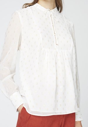Blouse blanche semi-transparente avec un motif subtil de carrés dorés, manches longues, devant froncé, lien au cou, portée avec un pantalon couleur rouille.