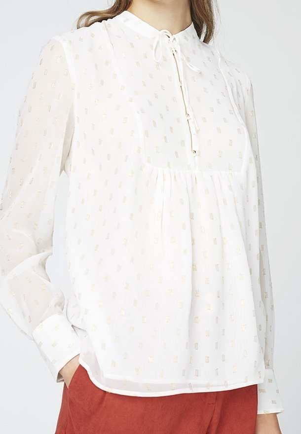 Blouse blanche semi-transparente avec un motif subtil de carrés dorés, manches longues, devant froncé, lien au cou, portée avec un pantalon couleur rouille.