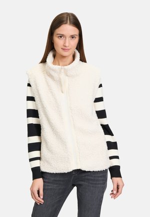 Gilet en polaire blanche avec col monté et fermeture éclair sur le devant, associé à une chemise à rayures noir et blanc à manches longues, modèle portant un jean gris.
