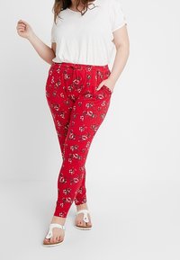 Žena v bílém tričku s krátkým rukávem, červených kalhotách s květinovým vzorem a kapsami, která stojí s jednou rukou v kapse.