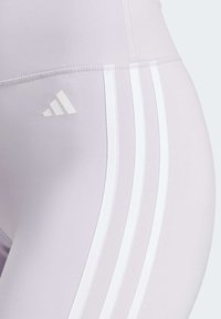 Lichtpaarse leggings met een witte drie-strepen patroon langs de zijkant en een reliëf witte Adidas-logo op de linker bovenbeen. Gladde, elastische stof.