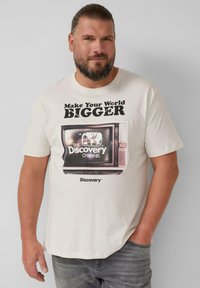 Lichtbeige katoenen t-shirt met een vintage tv-afbeelding. De tekst luidt "Maak je wereld GROTER" en "THE DISCOVERY CHANNEL" in vetgedrukte letters.