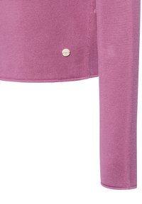 Close-up van de onderkant en de mouw van een textuur roze gebreide trui met een klein, rond metalen label dat vlakbij de zoom is bevestigd.