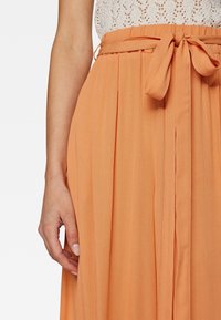 Perzikkleurige maxi jurk met een strik taille, met subtiele plooien en een lichte, gestructureerde stof. Close-up van een hand op de stof.