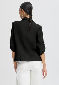 Giacca nera con colletto, maniche a tre quarti, tessuto leggermente strutturato e vestibilità blanda, abbinata a pantaloni bianchi.
