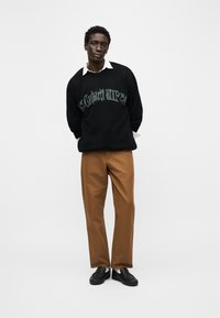 Άνδρας που στέκεται με τα χέρια πίσω από την πλάτη, φοράει μαύρο πουλόβερ με ξεθωριασμένο κείμενο, λευκό πουκάμισο με γιακά, καφέ παντελόνι και μαύρα μοκασίνια σε λευκό φόντο.