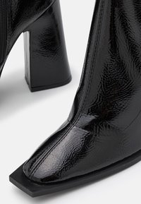 Bottine en cuir verni noir avec un bout carré, une texture brillante et un talon bloc épais. Dotée d'une fermeture éclair latérale.