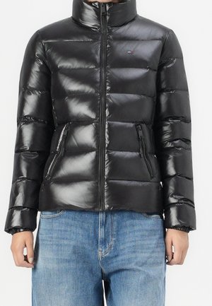 Chaqueta acolchada negra brillante con cuello alto y bolsillos con cremallera, combinada con vaqueros azules, vista frontal, manos relajadas a los lados.