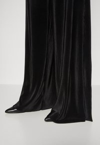 Černé, ribované, široké kalhoty vyrobené z hladké, lesklé tkaniny, doplněné o špičaté černé podpatky; zdůrazňují elegantní siluetu a texturu látky.