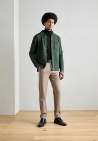 Chaqueta de punto verde con botones marrones, suéter de cuello alto negro, pantalones beige y zapatos negros. El modelo está de pie sobre un suelo de madera contra una pared clara.