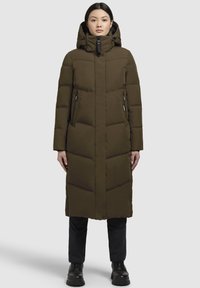  Lange olijfgroene gewatteerde jas met een capuchon, geruite uitstraling, zijzakken en een ritssluiting, gecombineerd met zwarte broek en laarzen.