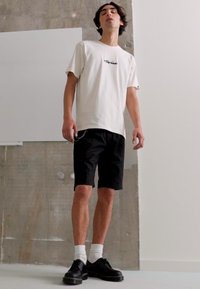 Weißes Grafik-T-Shirt, schwarze Shorts, schwarze Schuhe mit dicken Sohlen und weiße Socken. Einfaches Design, lässige Passform. Betonhintergrund.