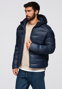 Giacca piumino navy con cappuccio, caratterizzata da un design trapuntato, chiusura a zip e polsini elasticizzati. Indossata sopra un maglione leggero e jeans di denim.