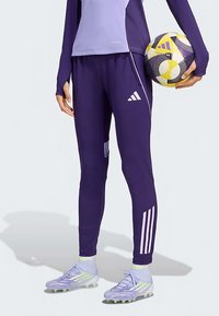 Person, der lila Adidas-Sportbekleidung trägt und einen gelb-lila Fußball hält, trägt Stollenschuhe mit neonfarbenen Akzenten.