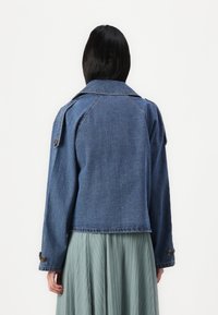 Temna modra denim jakna s strukturovanim ovratnikom, gumbi na manšetah in epauletti na ramenih; oblečena čez godrnjavo svetlo zeleno krilo.
