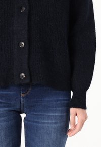 Svart stickad cardigan med mjuk textur, med runda knappar och avslappnad passform, i kombination med blå jeans. Enkel bakgrund.