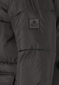 Svart vadderad jacka med en glänsande yta, horisontalt quiltade sektioner och en fyrkantig logotyp patch på övre ärmen.