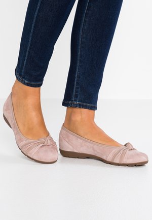 Des ballerines en daim rose présentent un détail de nœud tordu au niveau des orteils et une semelle marron flexible, qui se marient bien avec un jean en denim.
