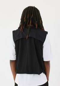 Sort ærmeløs vest med bred krave over en hvid t-shirt. Glat stof med en subtil tekstur, set fra ryggen mod en ensfarvet baggrund.