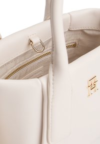 Λευκή δερμάτινη τσάντα χειρός με χρυσά μεταλλικά στοιχεία, λαβές στο επάνω μέρος και κλείσιμο με φερμουάρ. Το εσωτερικό έχει επενδυμένο διαμέρισμα και λεπτομέρεια με λογότυπο.